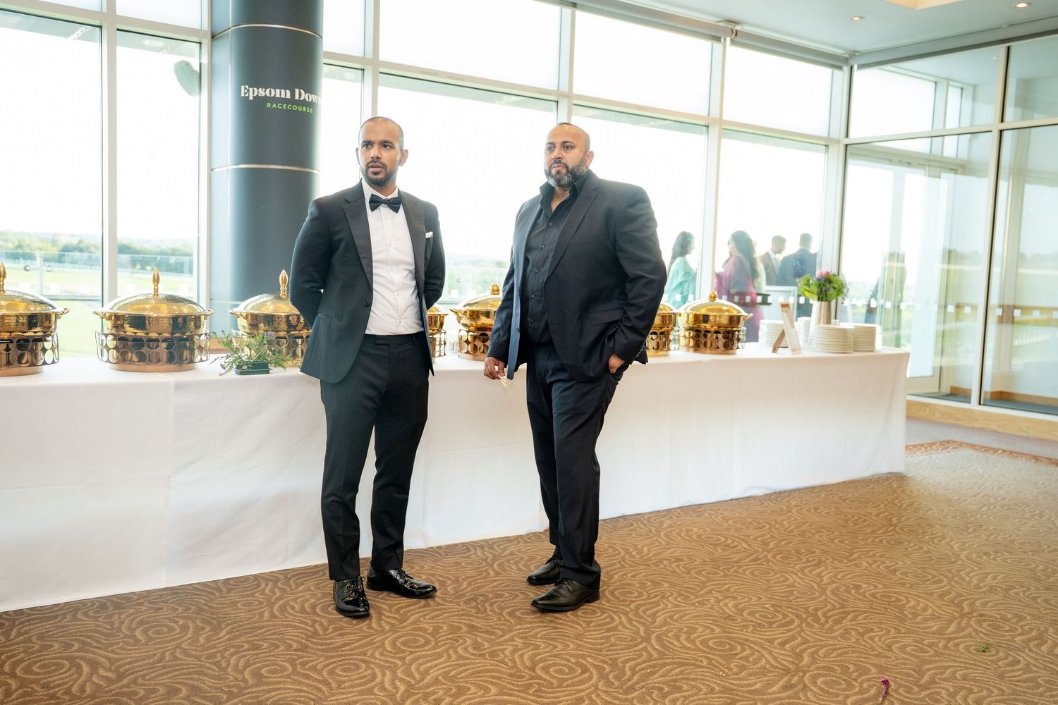 Two well-dressed men in dark suits posing together at a formal venue with large windows.