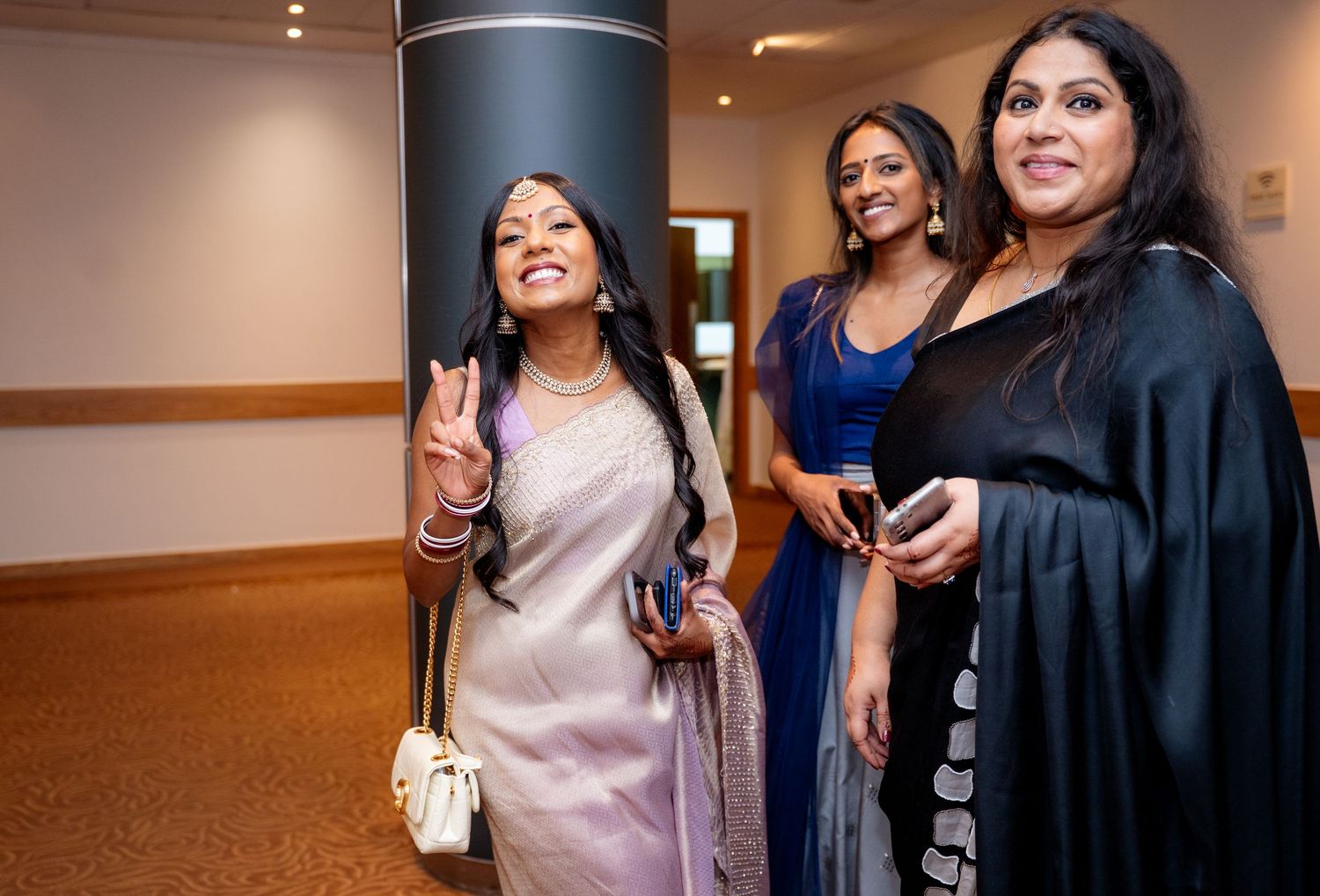 Three women in elegant attire at an indoor event, smiling and socializing in a modern venue with ambient lighting.
