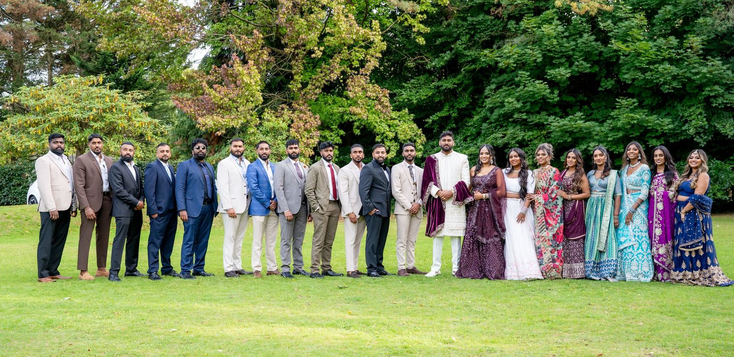 Large wedding party lineup in traditional Indian and formal attire posing on a green lawn surrounded by trees.