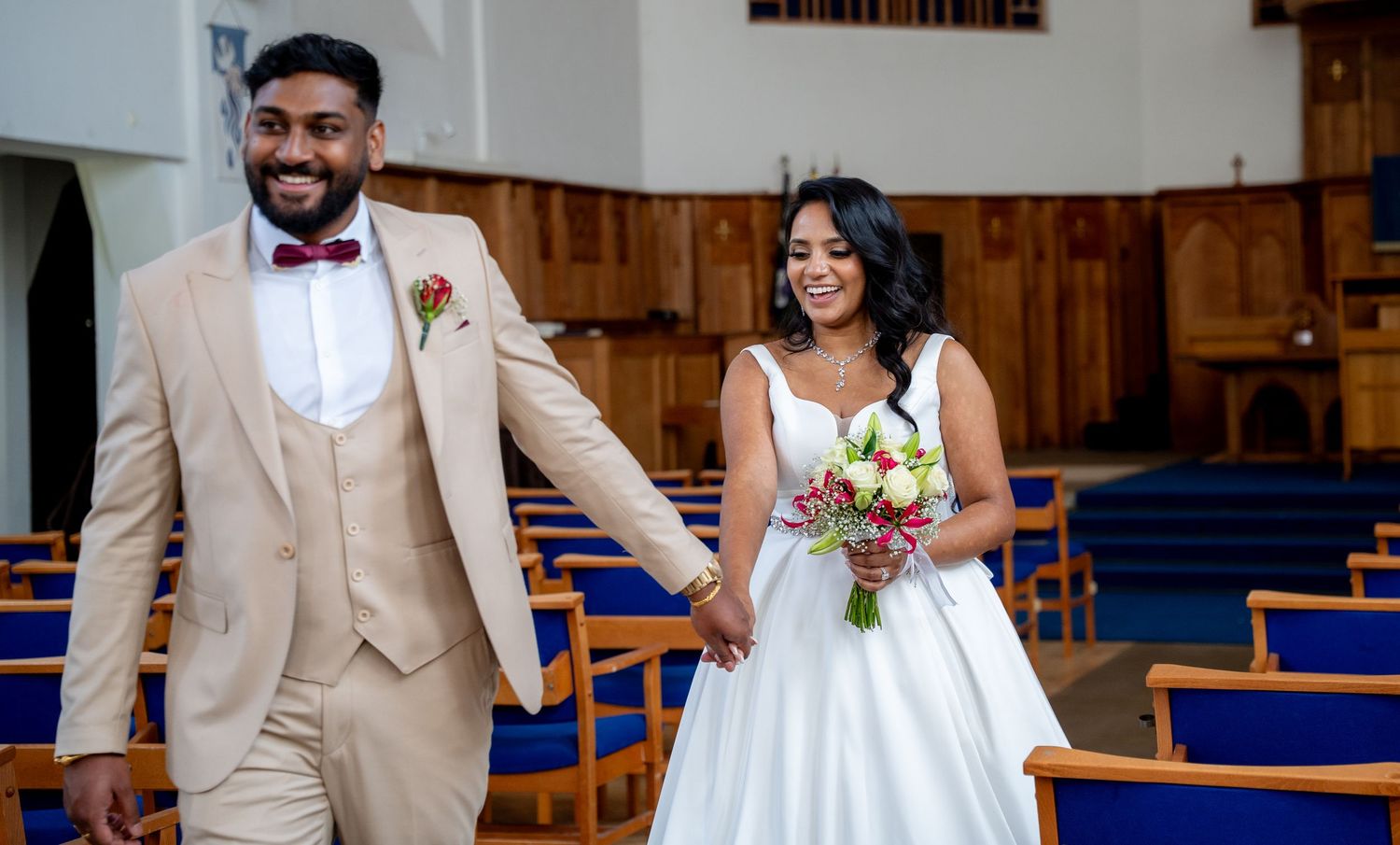 Bride and groom walking hand in hand down church aisle, groom in tan suit, bride in white gown holding bouquet.