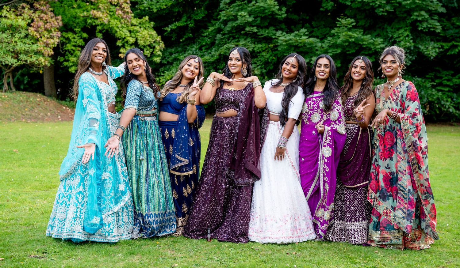 Group of women in vibrant Indian traditional attire posing outdoors on green grass, wearing colorful lehengas and sarees.