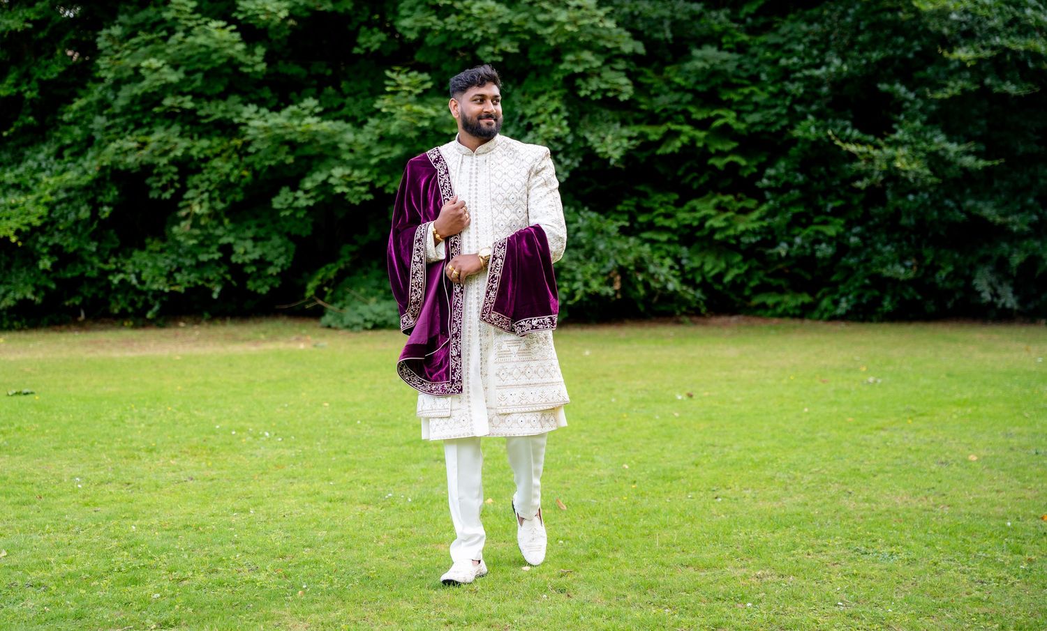 Indian groom in white embroidered sherwani with purple velvet shawl smiling and walking on green lawn at wedding.