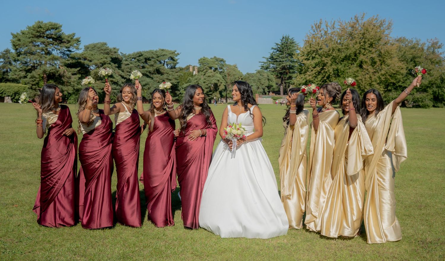 Bride with bouquet poses with bridesmaids in burgundy and gold gowns celebrating outdoors on sunny day.
