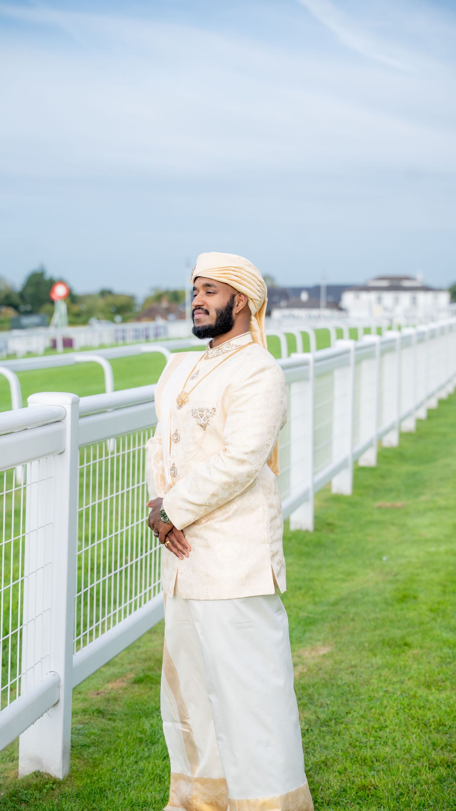 Man in traditional South Asian cream sherwani and turban standing by white racetrack fence on sunny day.