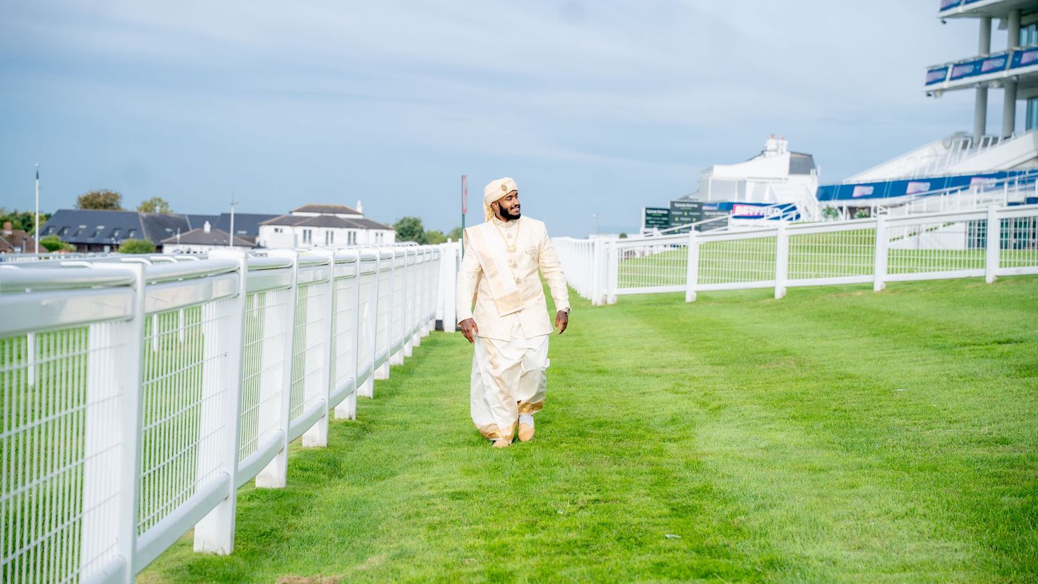Man in traditional South Asian attire walking on lush green grass at a racecourse with white fencing.