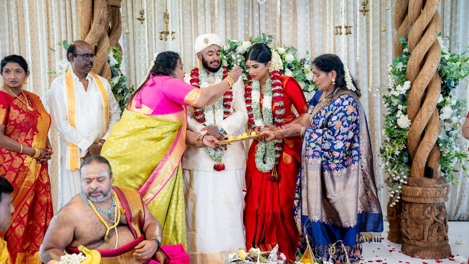 Traditional Indian wedding ceremony with bride in red silk saree and groom exchanging floral garlands with family present.