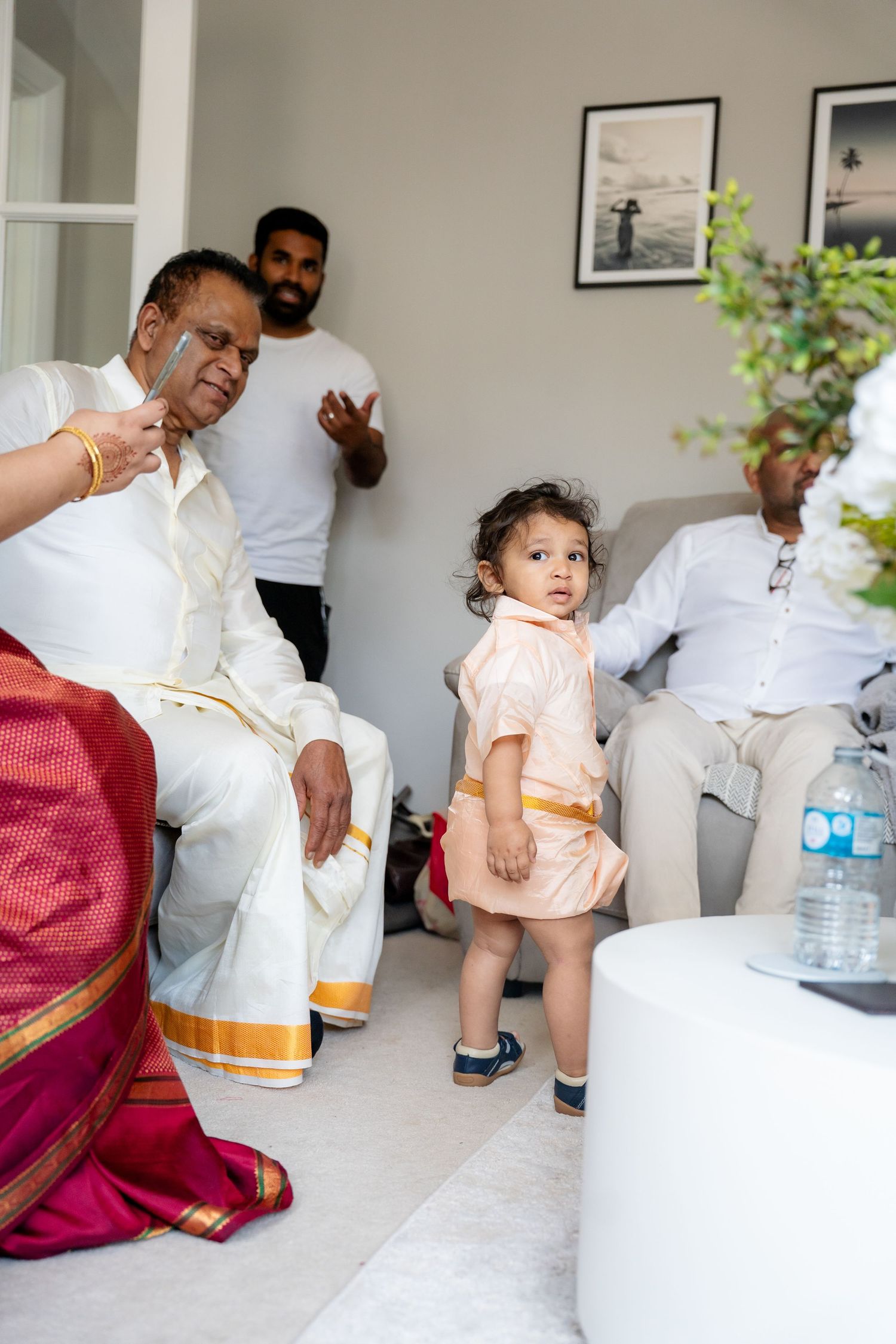 Young child in traditional peach outfit standing in living room during Indian wedding family gathering.