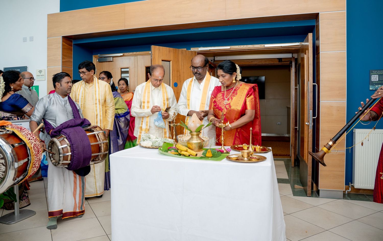 Group of people in traditional Indian attire perform a Hindu ritual ceremony with brass lamps and offerings indoors.