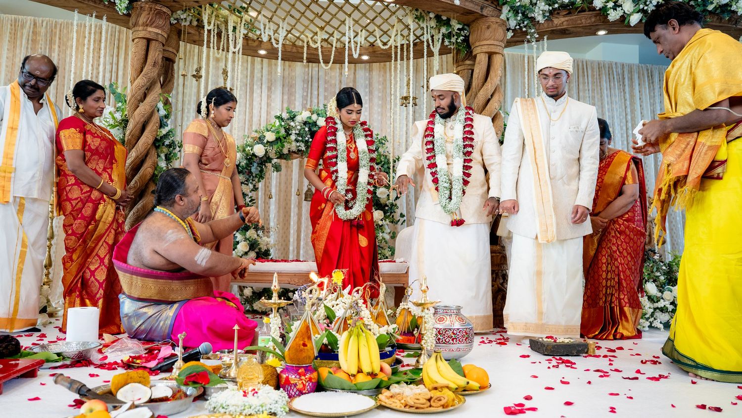 Traditional Hindu wedding ceremony with priest conducting rituals as bride in red and groom in white stand on flower-adorned stage.
