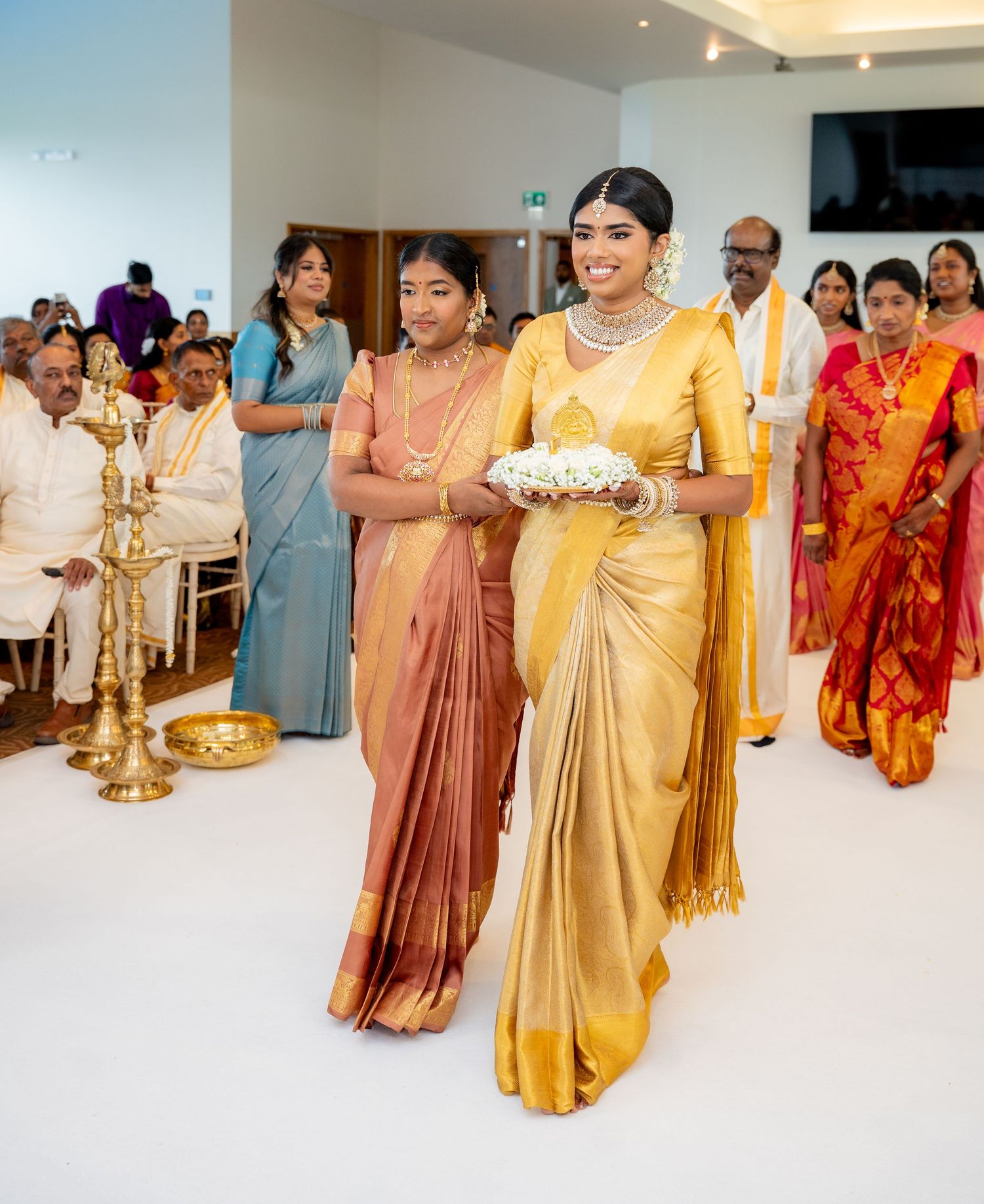 A bride in a gold silk saree carries flowers during a South Indian wedding ceremony with guests in traditional attire.