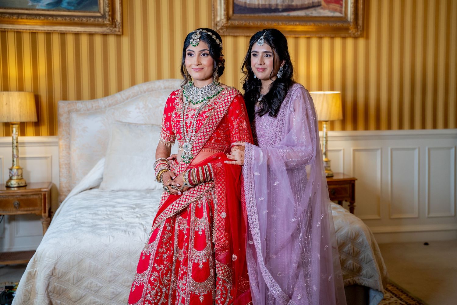 Indian bride in red embroidered lehenga poses with bridesmaid in lavender outfit on white bed.