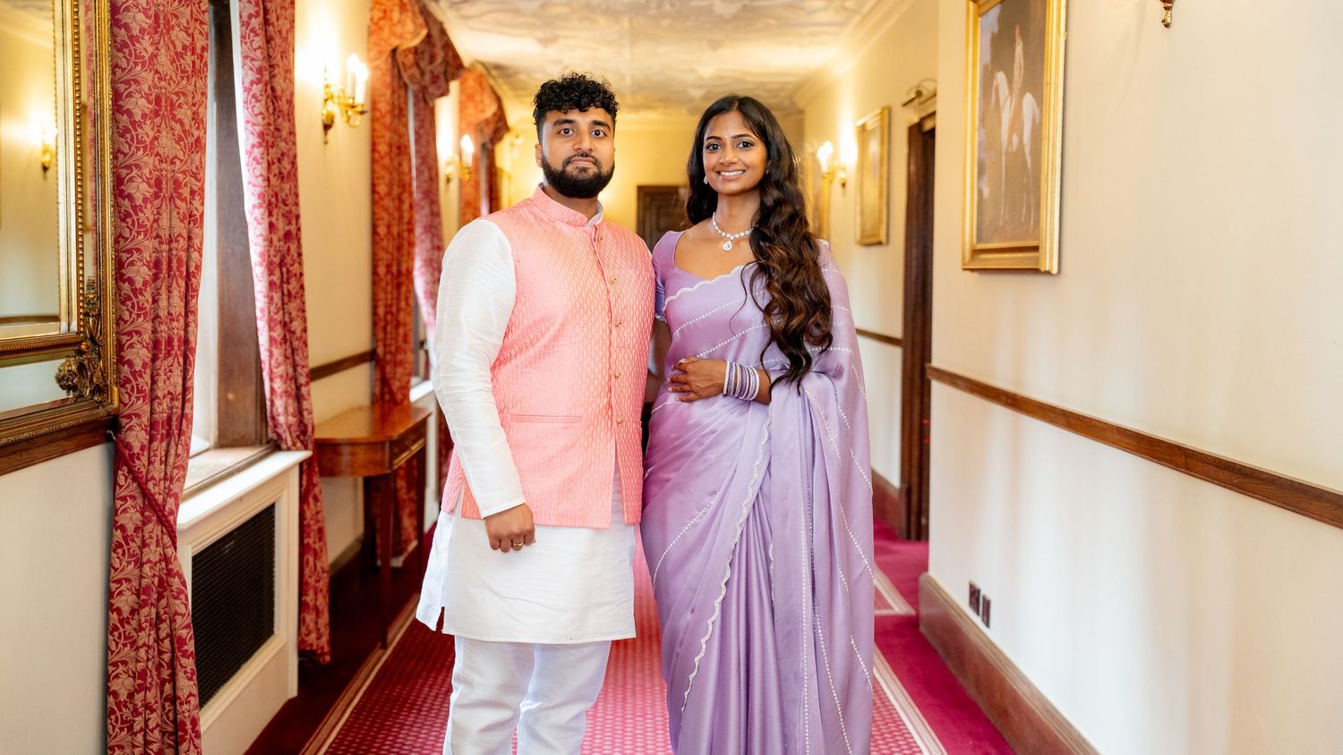 South Asian couple in coordinating lavender and pink wedding outfits posing in an elegant hallway.