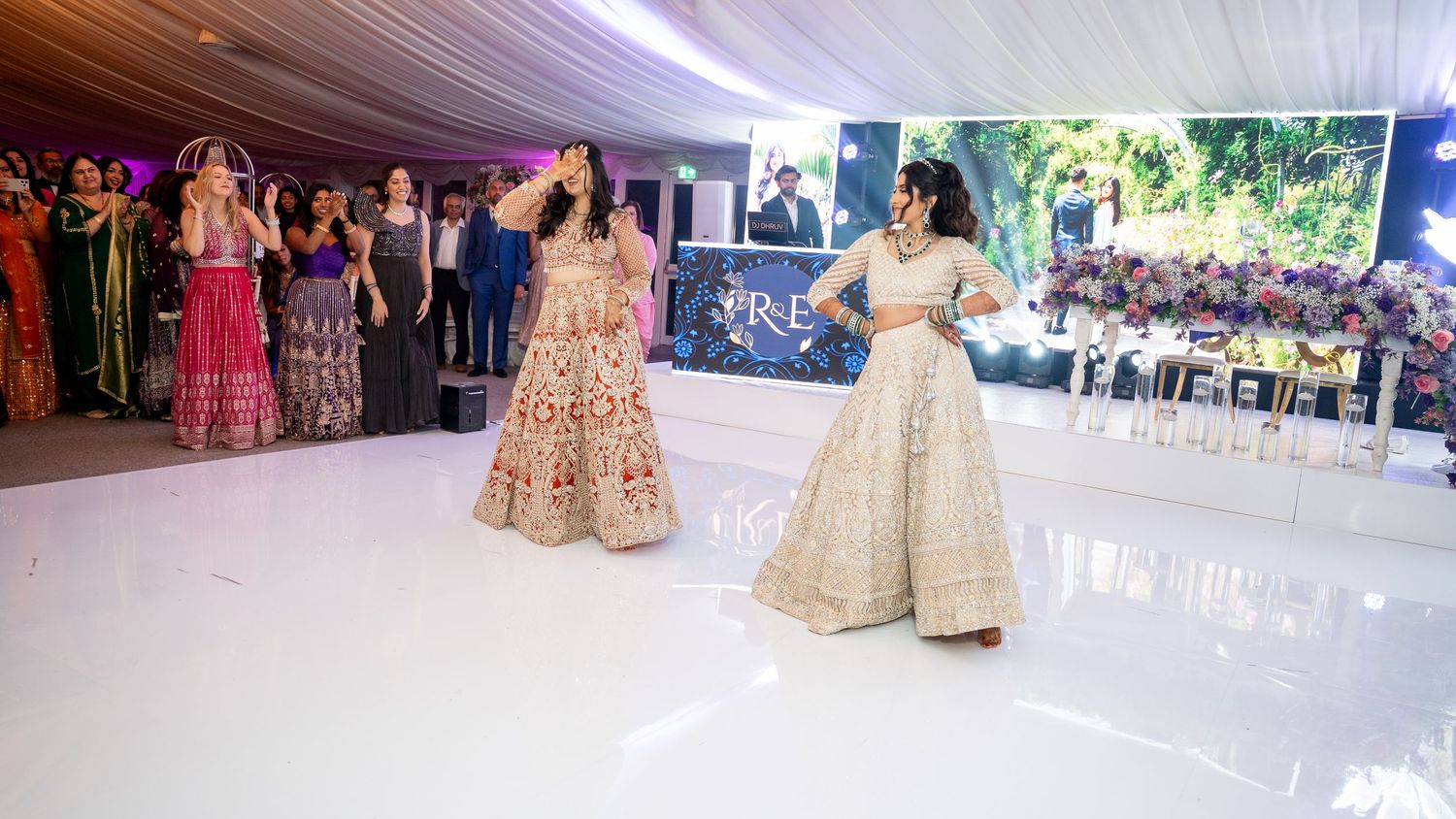 Two women in elegant Indian lehengas perform a dance at a wedding reception on a white dance floor.