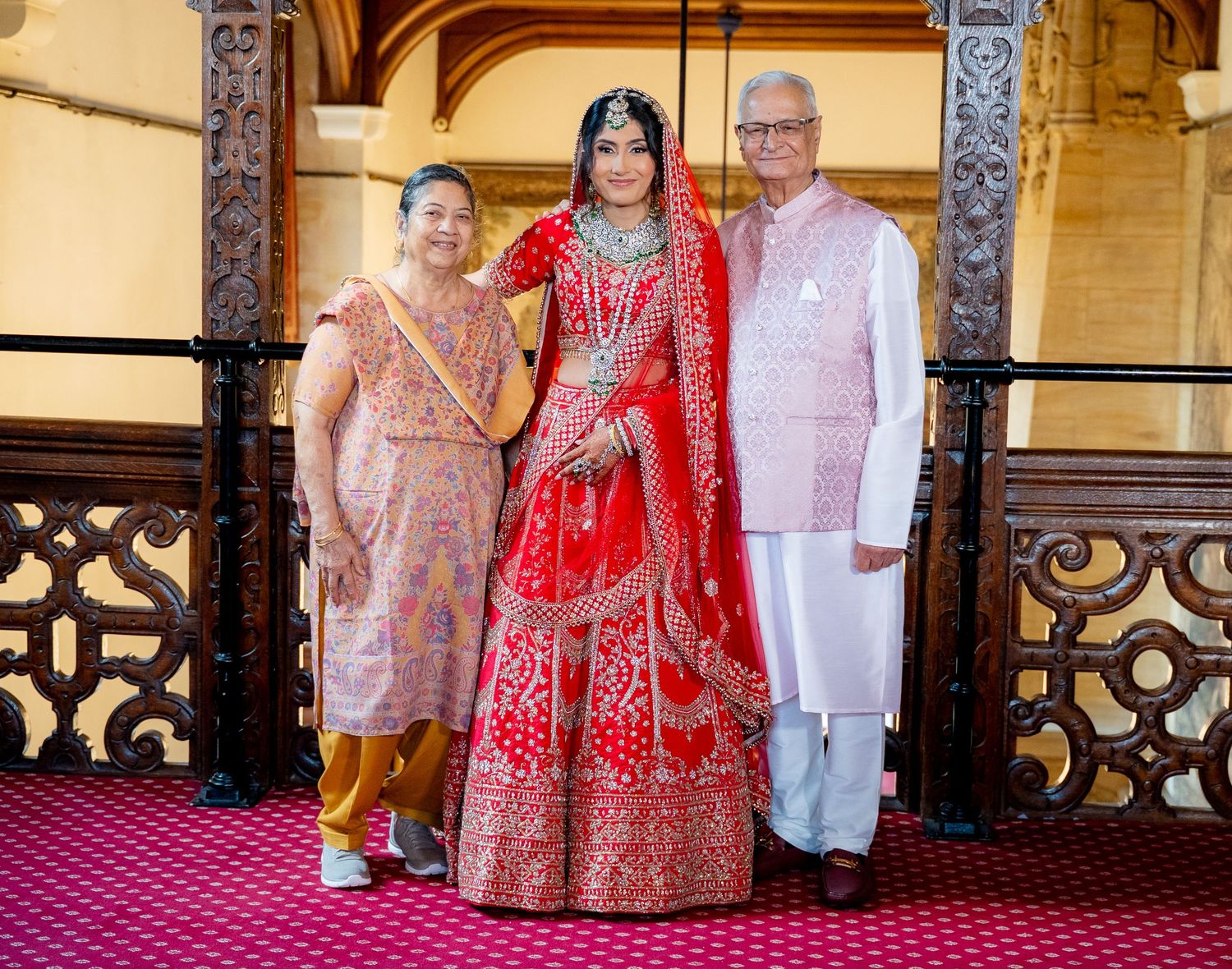 Indian bride in red lehenga poses with two family members on an ornate balcony.