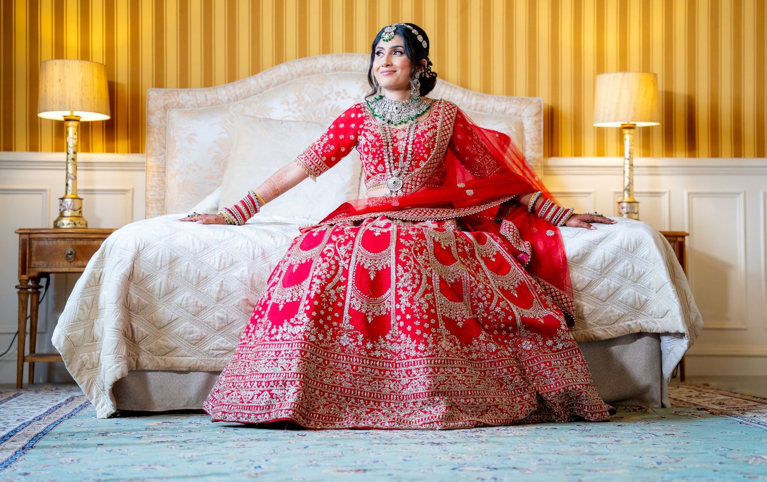 Indian bride in red embroidered lehenga sitting on white bed with gold lamps in elegant room.