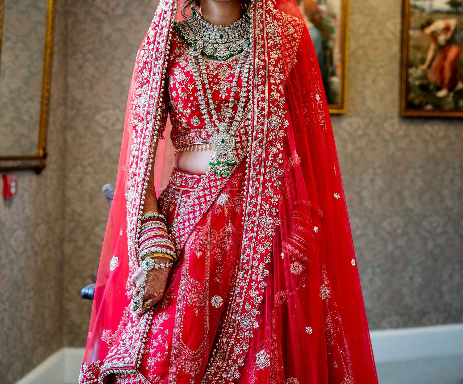 Indian bride in ornate red lehenga with gold embroidery, wearing traditional bridal jewelry and dupatta indoors.