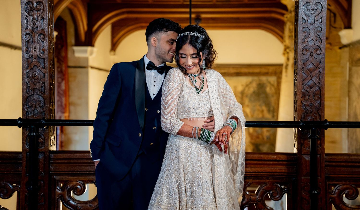 South Asian couple in elegant wedding attire pose on ornate staircase, bride in white lehenga, groom in navy suit.