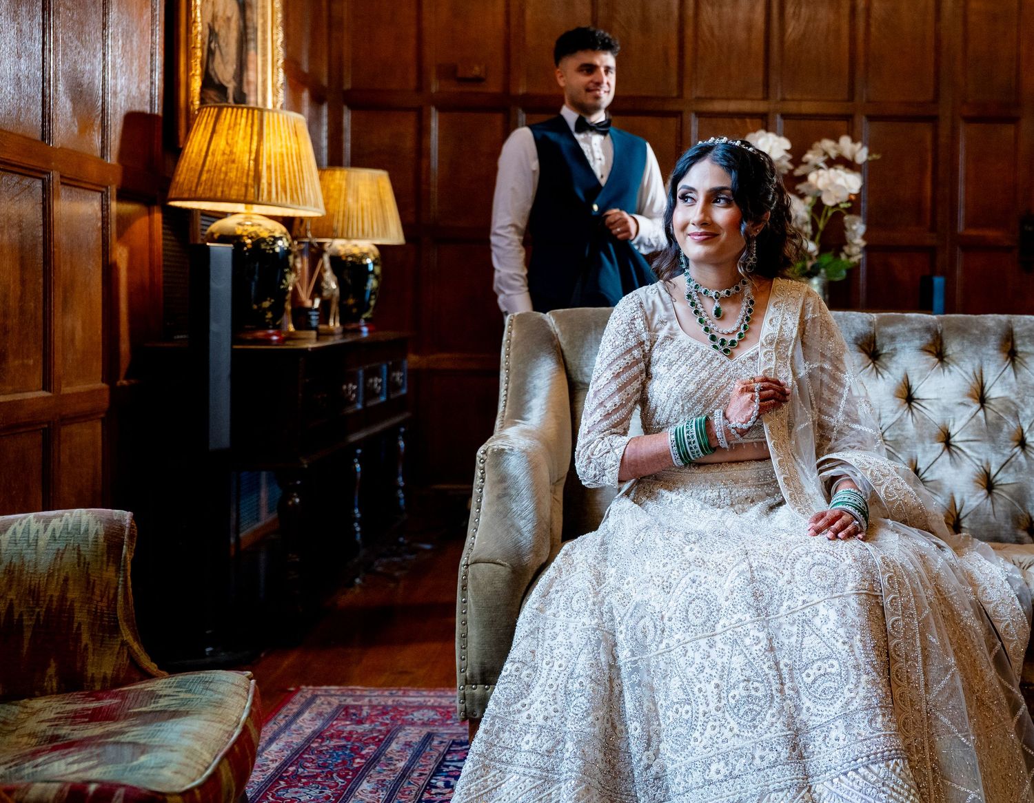 Bride in white embroidered lehenga sits elegantly in armchair while groom stands in background at wedding.