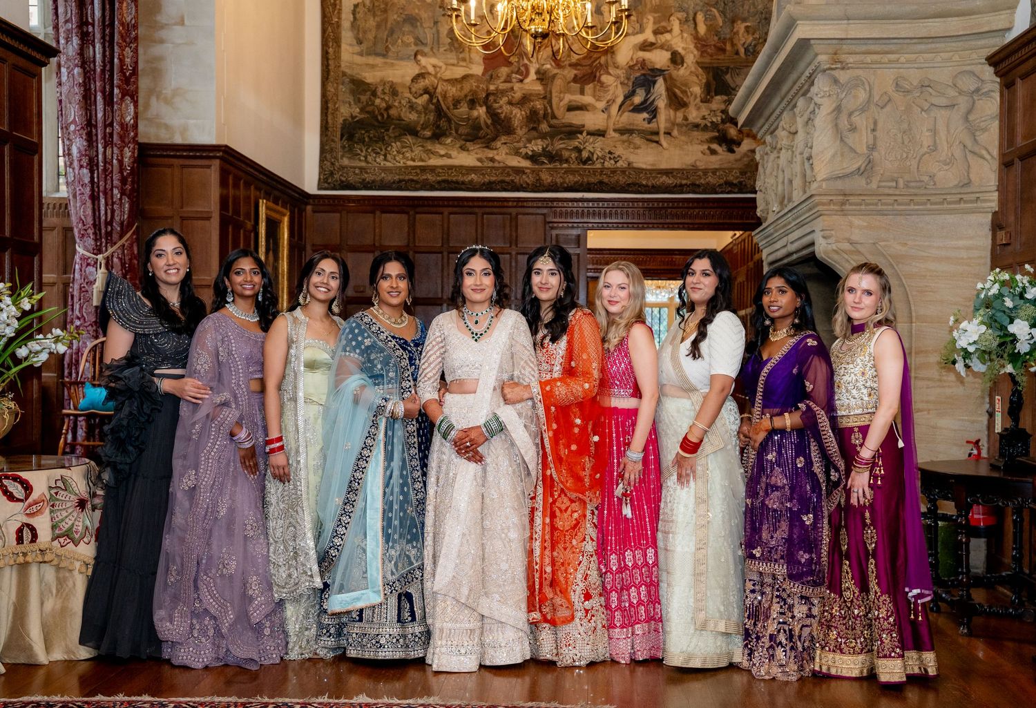Group of women in colorful Indian lehengas and sarees posing together in ornate ballroom setting.