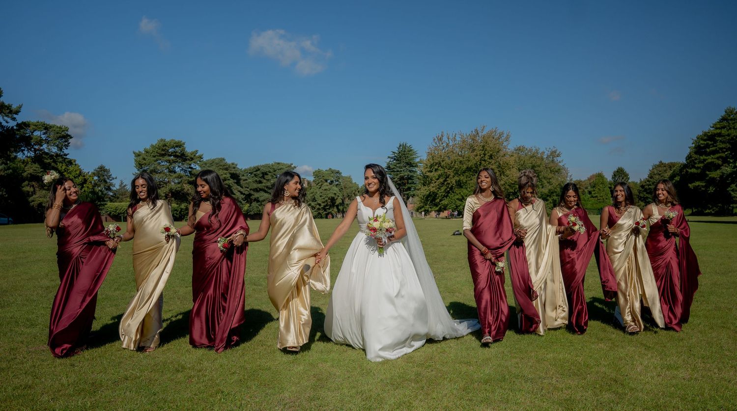 Bride in white wedding dress stands with bridesmaids in burgundy and gold gowns on sunny green lawn.