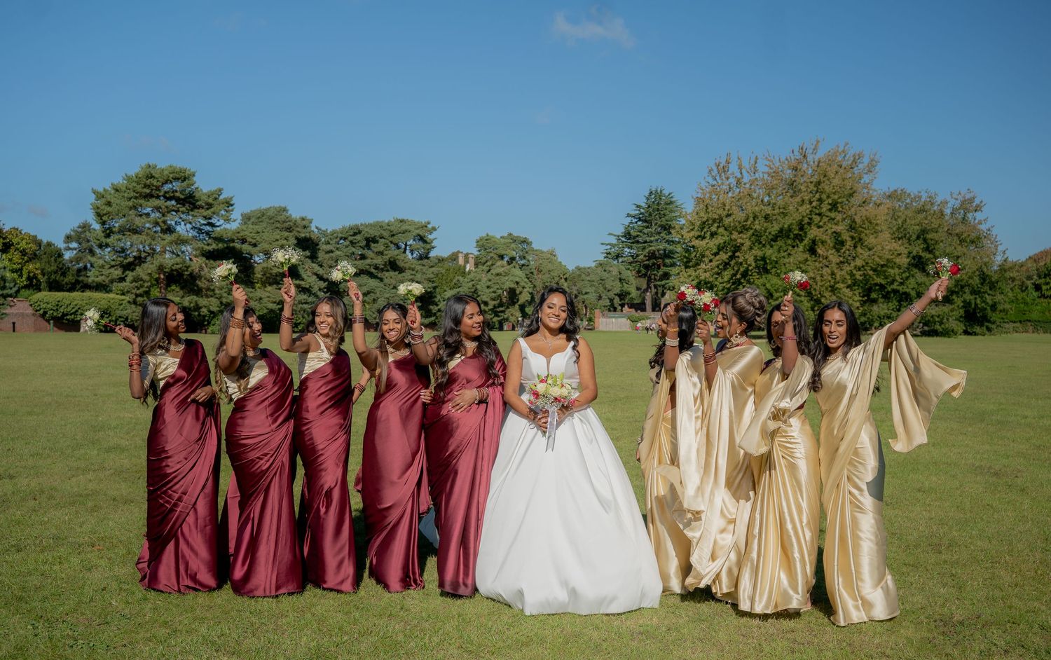 Bridesmaids in burgundy and gold sarees celebrate joyfully with bride on a sunny green lawn.
