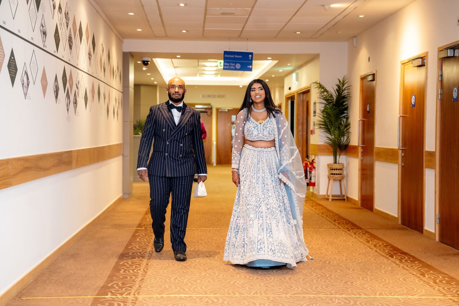 Couple in elegant Indian wedding attire walking down a decorated hotel corridor, man in navy suit, woman in silver lehenga.
