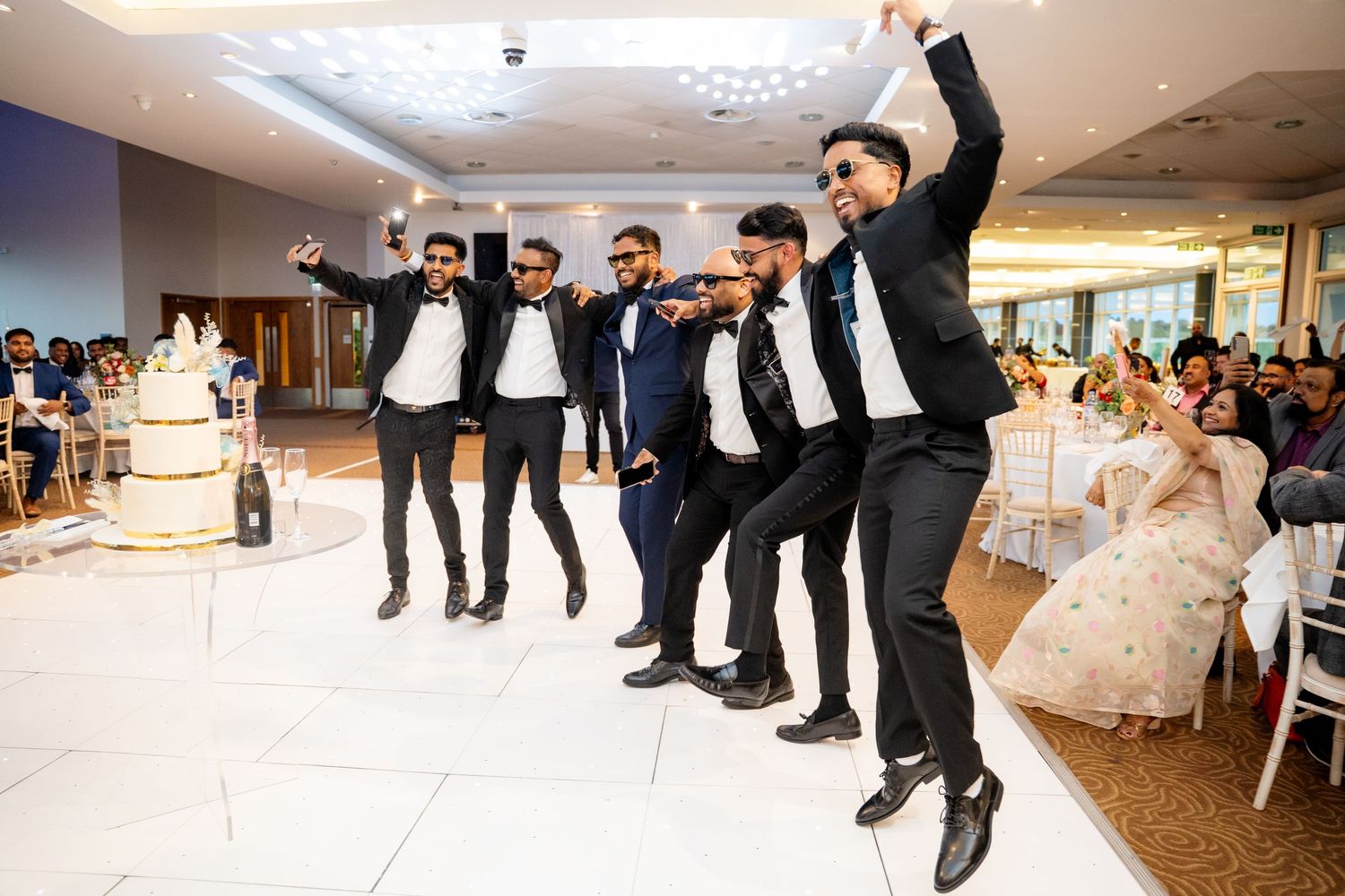Groomsmen in black suits performing a choreographed dance near the wedding cake as bride watches.