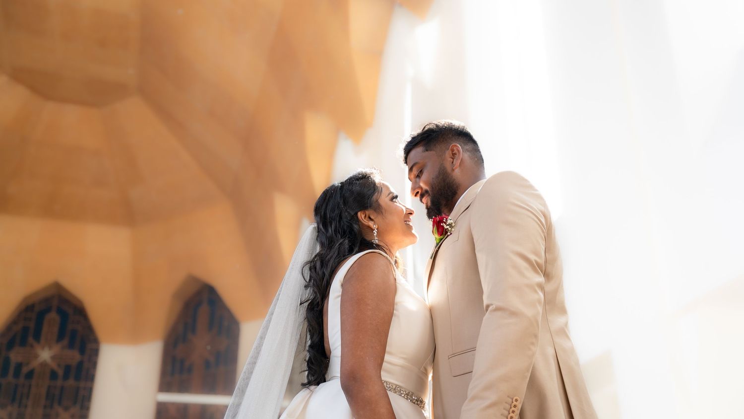 A bride and groom share a romantic moment outside a white church, with the bride in a veil and groom in a beige suit.