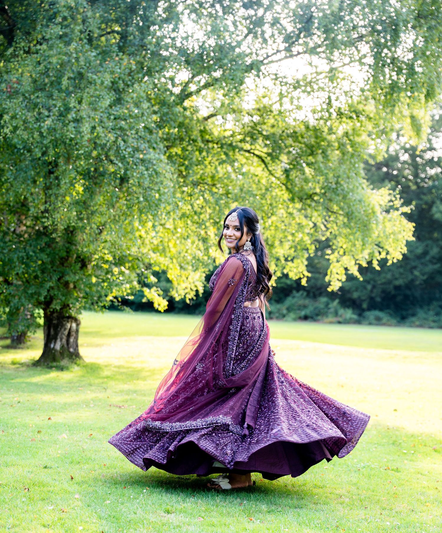 Woman twirling in a stunning purple embroidered lehenga on green grass with lush trees in the background.