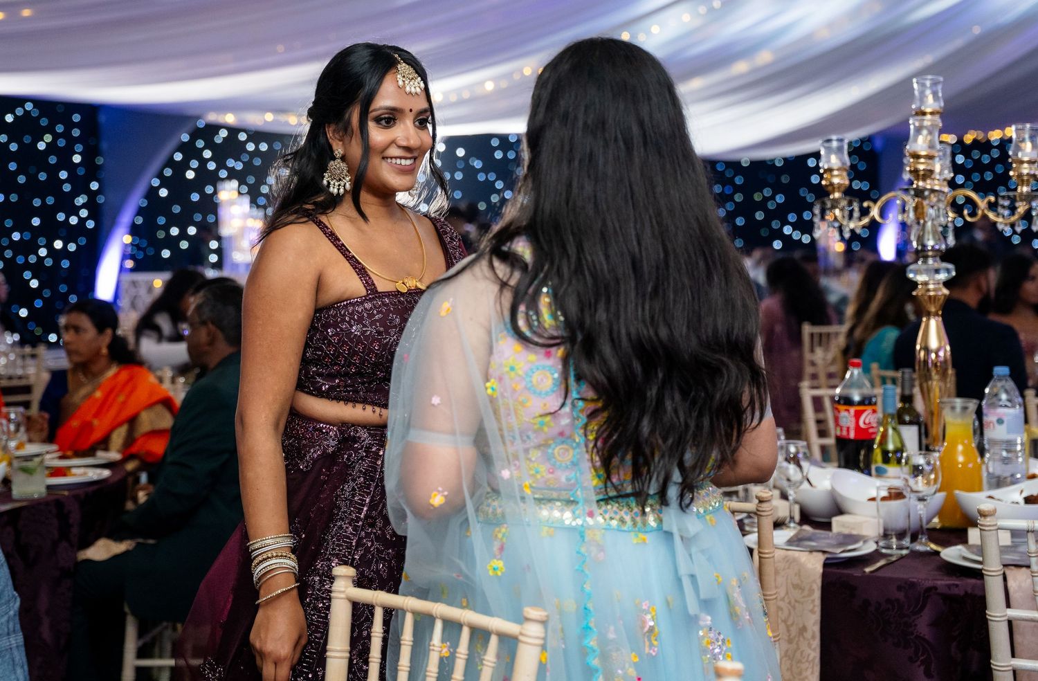Two women in colorful lehengas chat warmly at an elegantly decorated Indian wedding reception venue.
