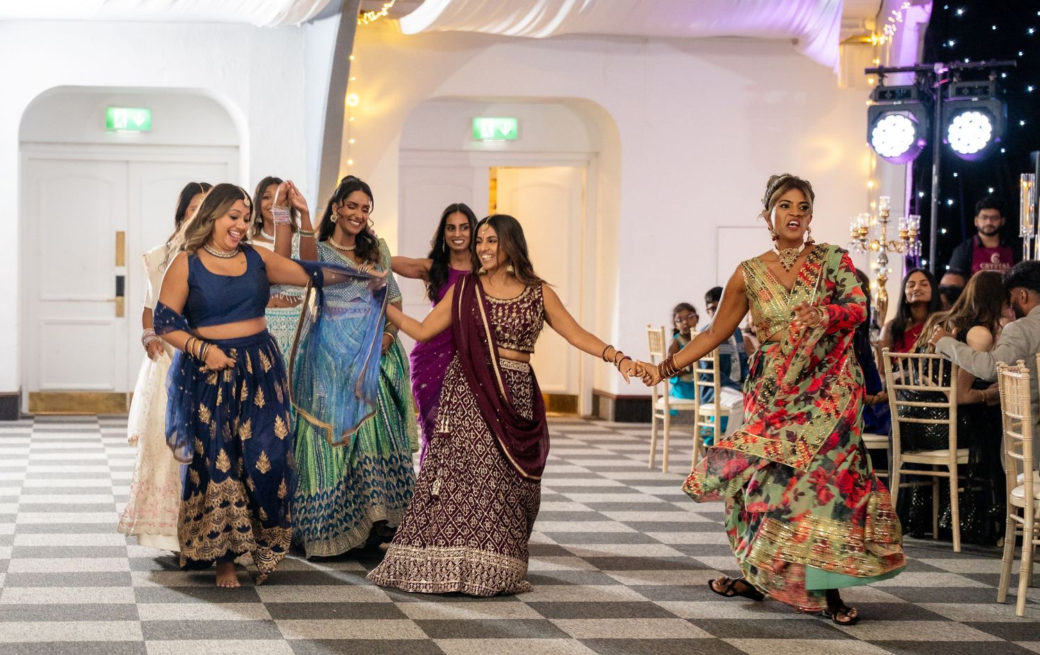 Bridesmaids in vibrant Indian attire holding hands and walking across a checkered dance floor.