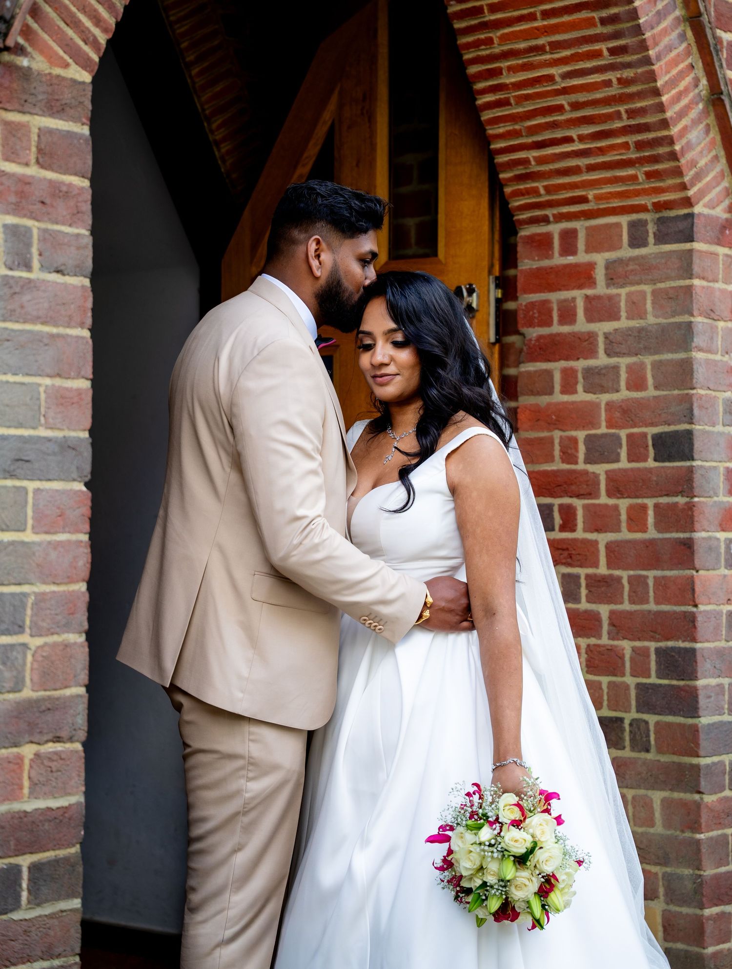 Bride in white gown holding bouquet and groom in beige suit embrace at brick church doorway.