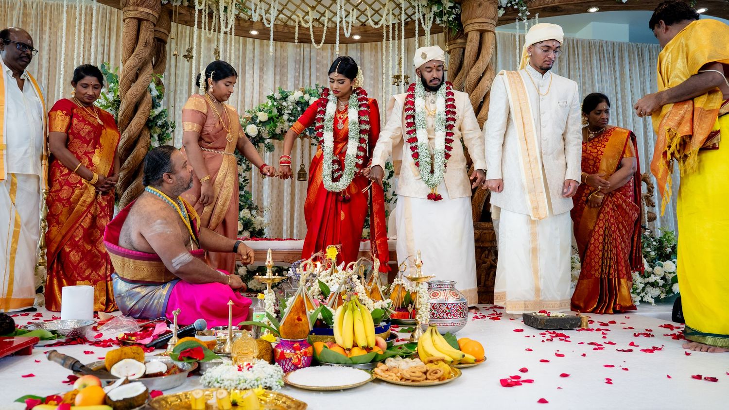 Colorful South Indian wedding ceremony with bride in red saree and groom in white standing before sacred ritual offerings.