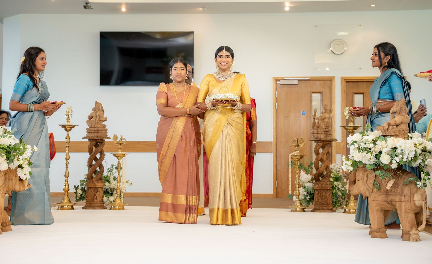 Two women in colorful silk sarees stand at a traditional South Indian wedding ceremony with brass lamps and floral decor.
