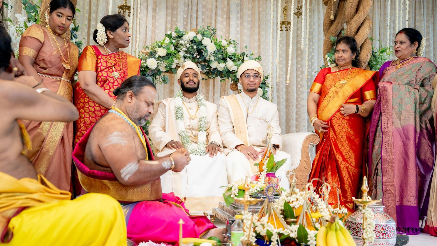 Two grooms seated at a South Indian wedding mandap with priest and family in colorful sarees.