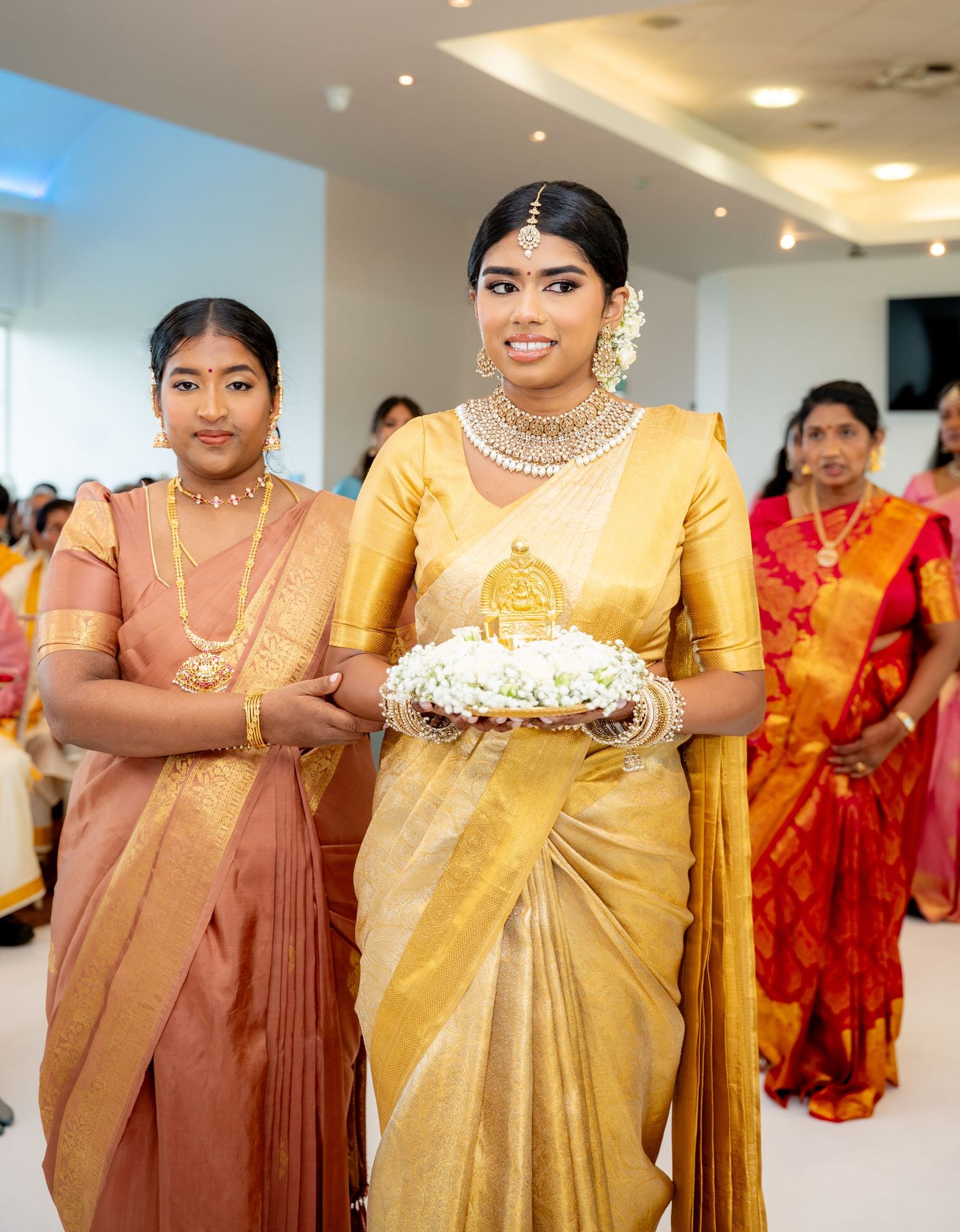 Two women in traditional South Indian gold and pink silk sarees walk in a ceremonial procession holding a floral tray.