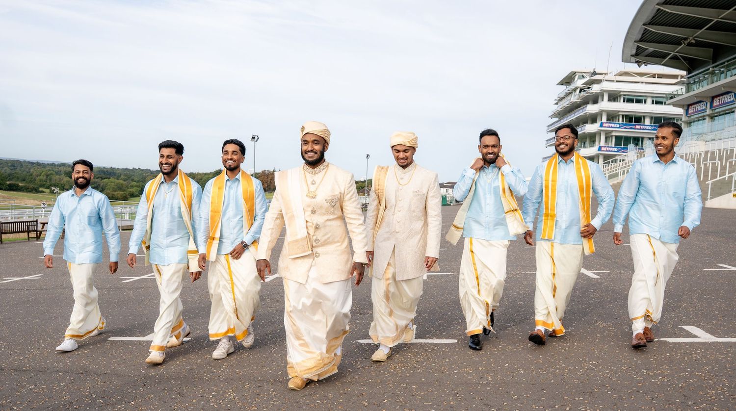 Groom and groomsmen in traditional Indian attire walking confidently outdoors near a racetrack on a wedding day.