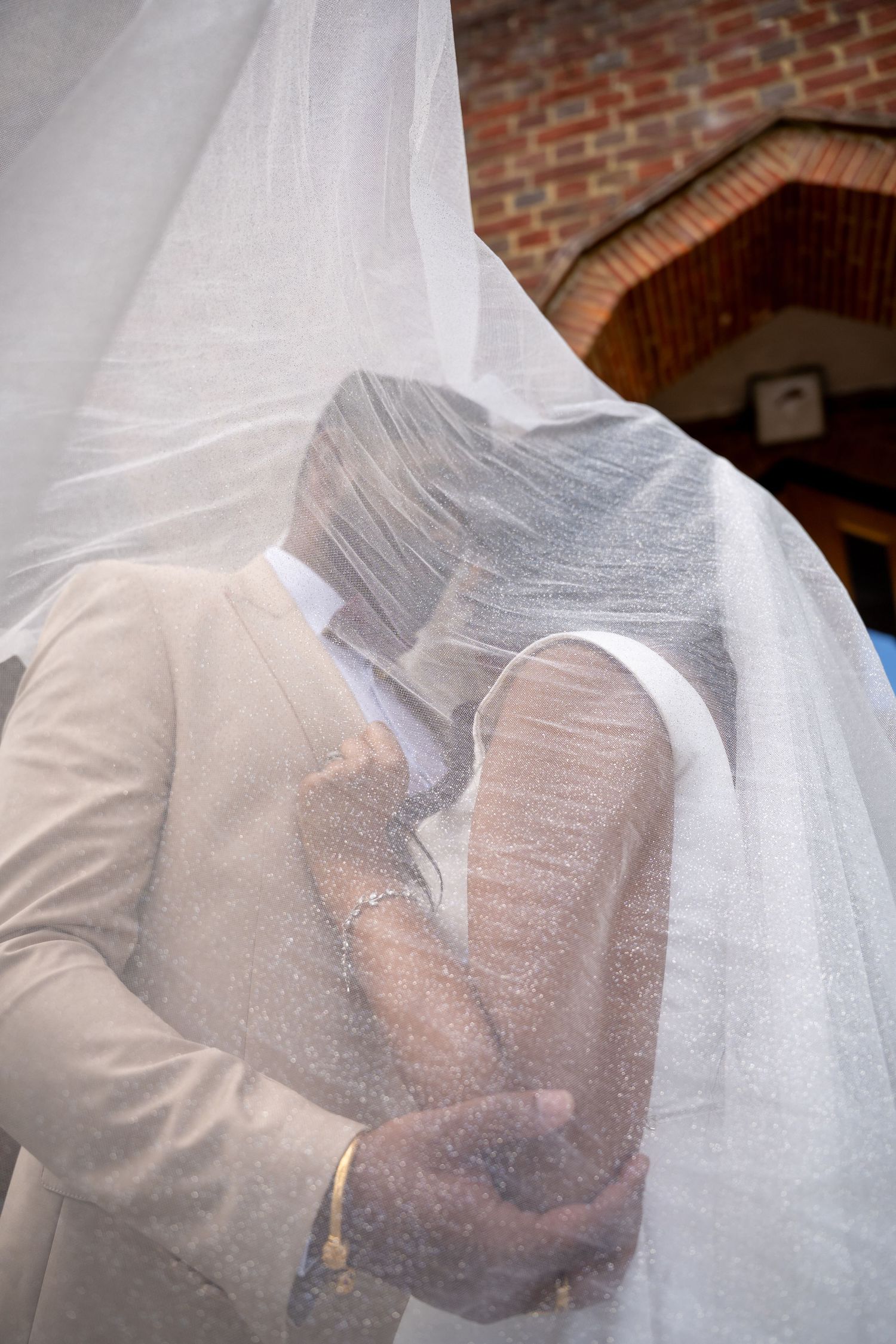 Bride and groom sharing a romantic kiss under a flowing white veil outside a brick church on their wedding day.