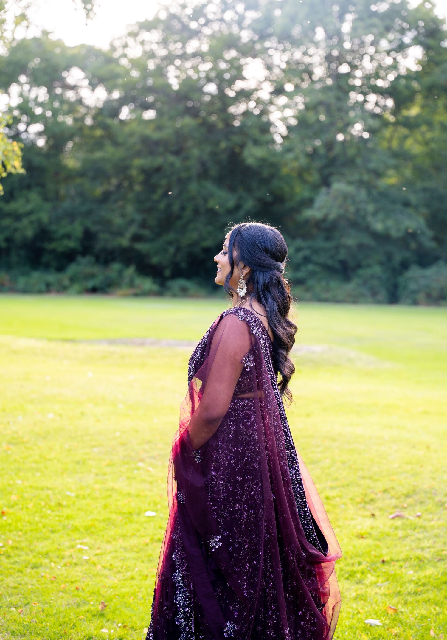 A bride in a stunning purple embellished lehenga stands on lush green grass, showcasing her elegant half-up hairstyle.