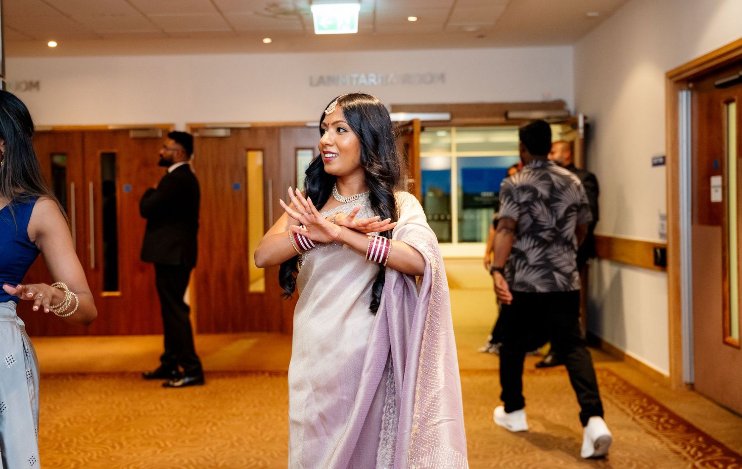 Woman in lavender saree gesturing expressively in a formal event venue corridor.