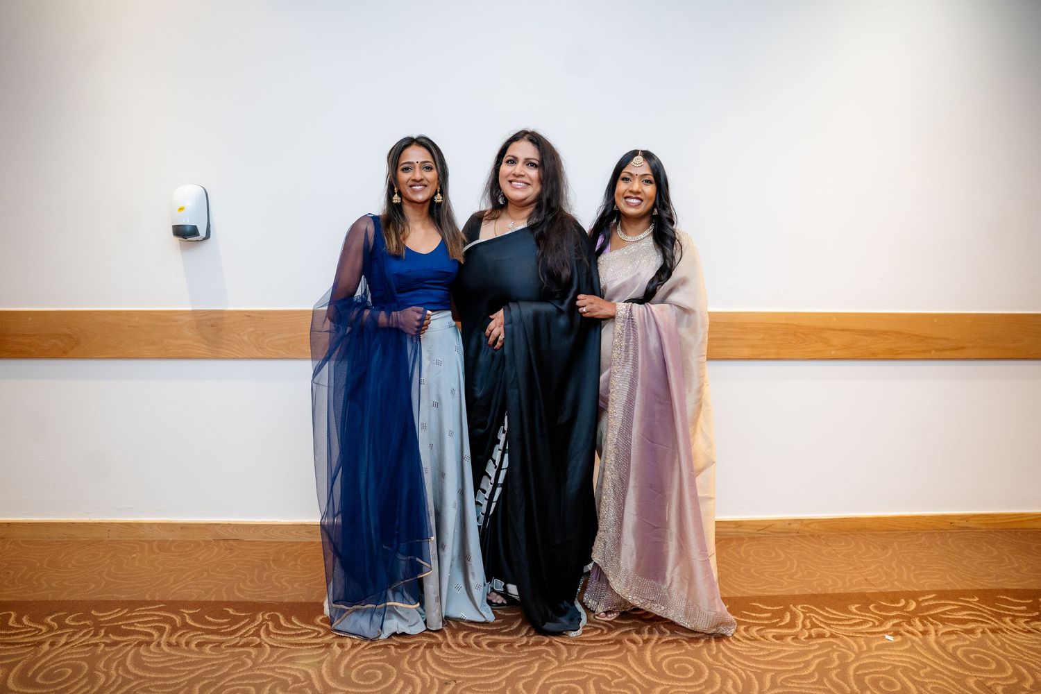 Three women in elegant Indian attire pose together smiling at a formal event.