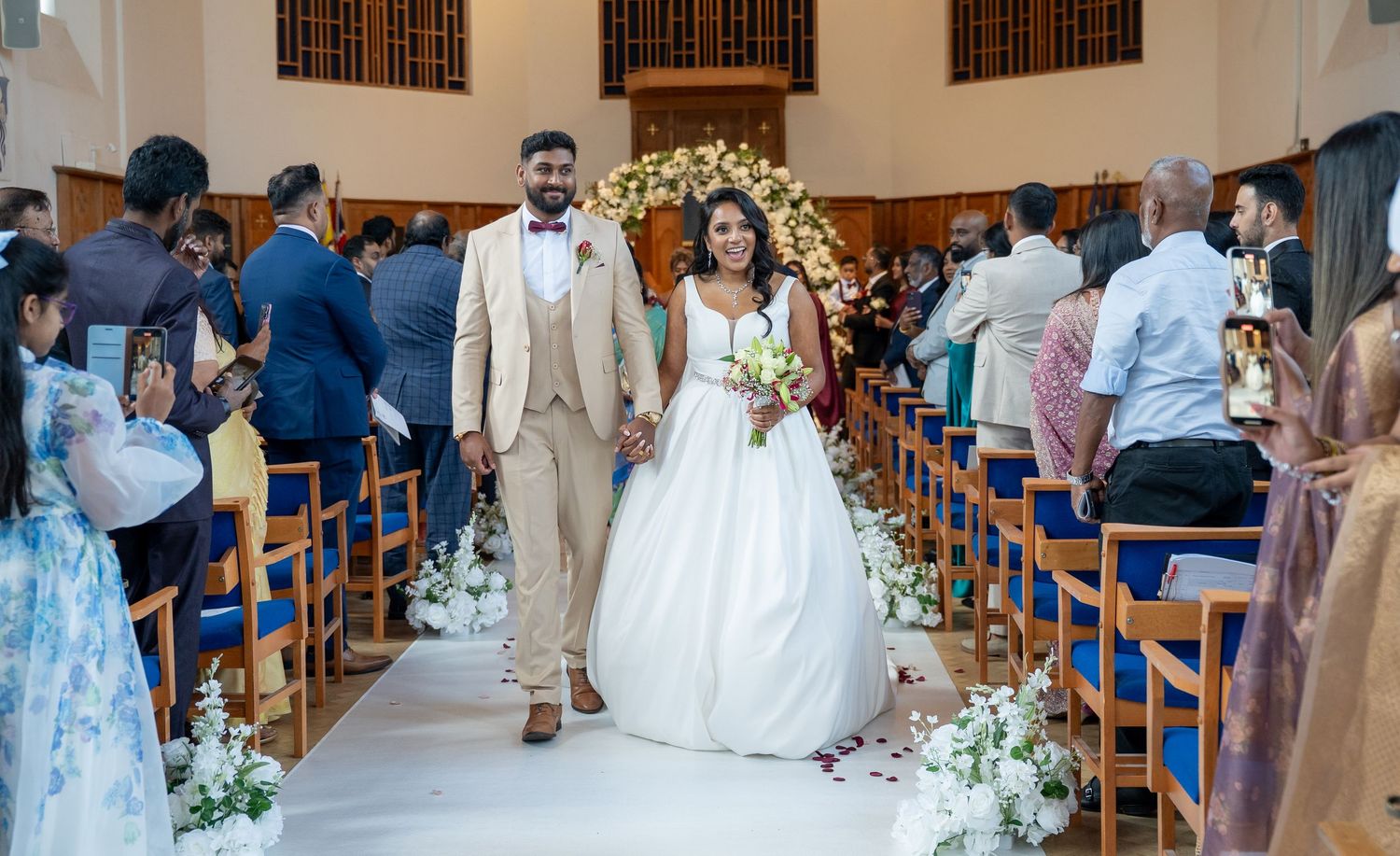 Happy bride and groom walk down church aisle holding hands, guests applaud in decorated pews around them.