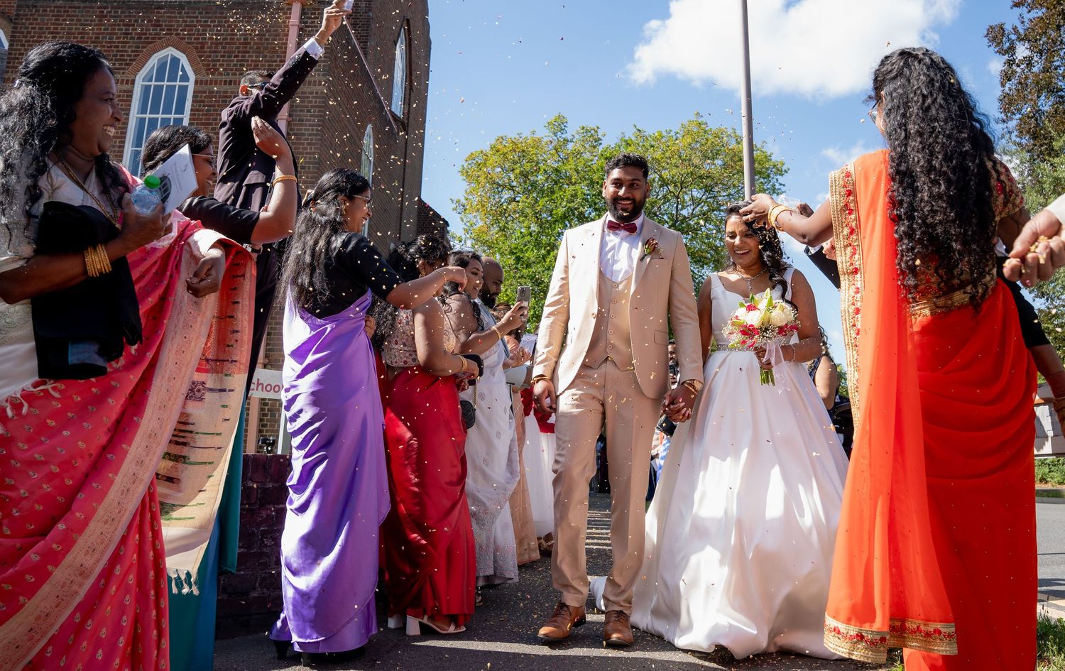 Newlyweds walk through a colorful confetti celebration surrounded by guests in vibrant traditional attire.