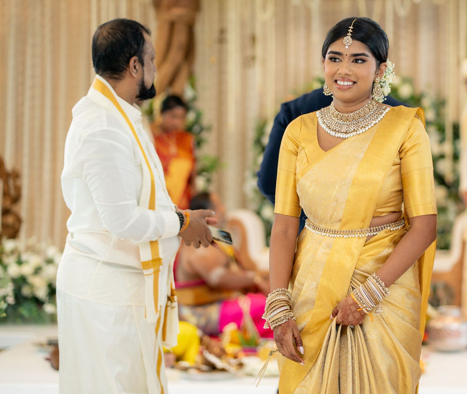 Bride in golden yellow silk saree with diamond jewelry smiling at groom during South Indian wedding ceremony.