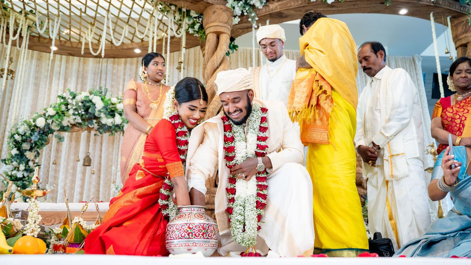 Bride and groom perform a traditional water pot ritual surrounded by family at a South Indian wedding.