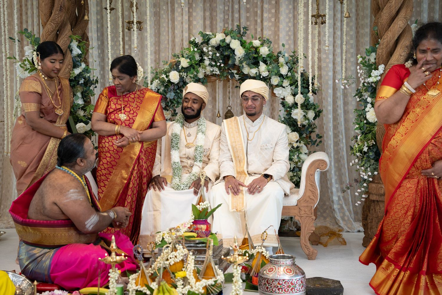 Two grooms seated at a floral South Indian wedding mandap during traditional Hindu ceremony.