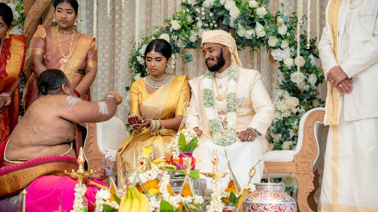 Couple in traditional South Indian wedding attire seated at decorated altar with priest and guests during ceremony.