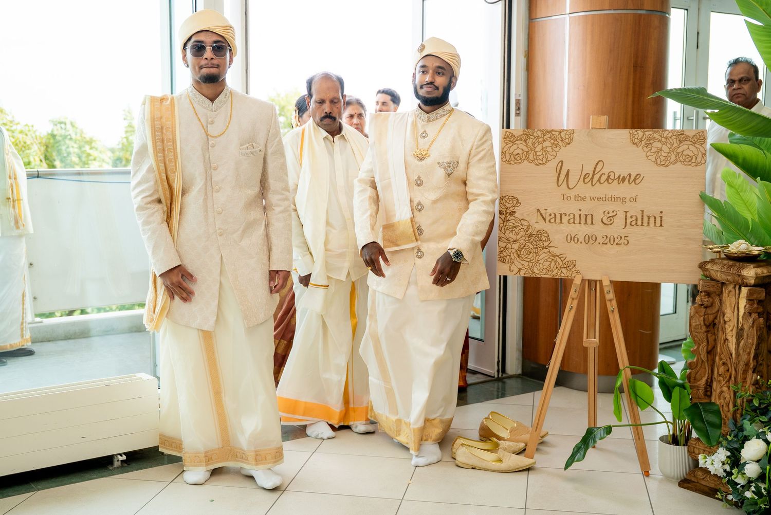 Two grooms in white Indian wedding attire stand beside a welcome sign for Narain and Jaini wedding.