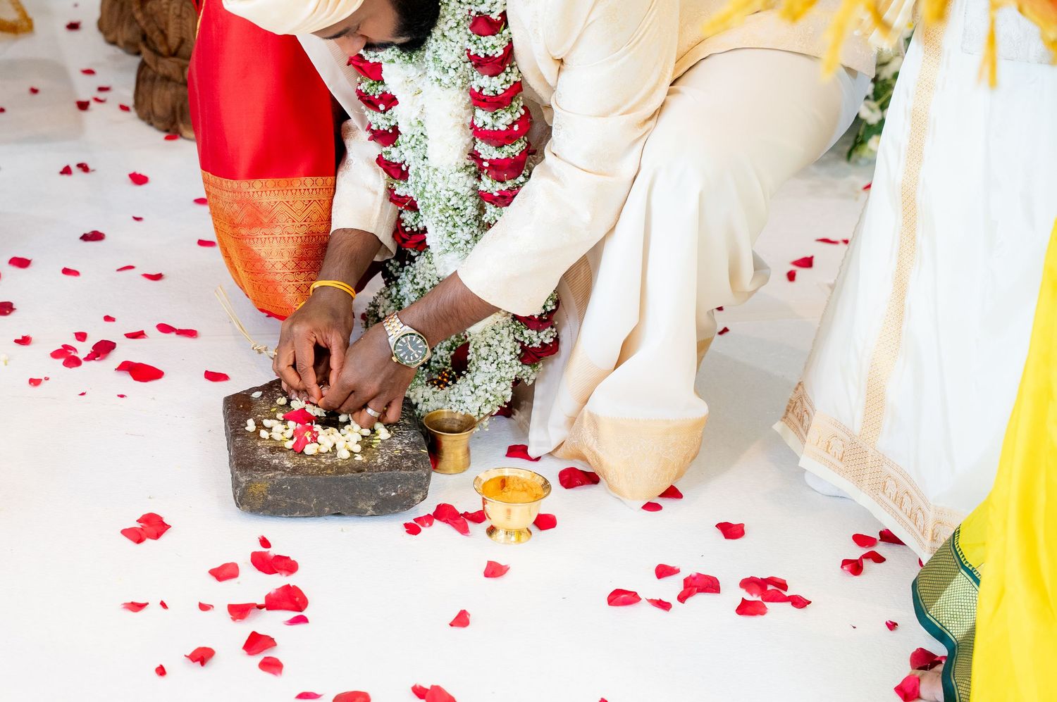 Close-up of South Indian wedding ritual showing couple placing flowers on sacred stone amid scattered red rose petals.