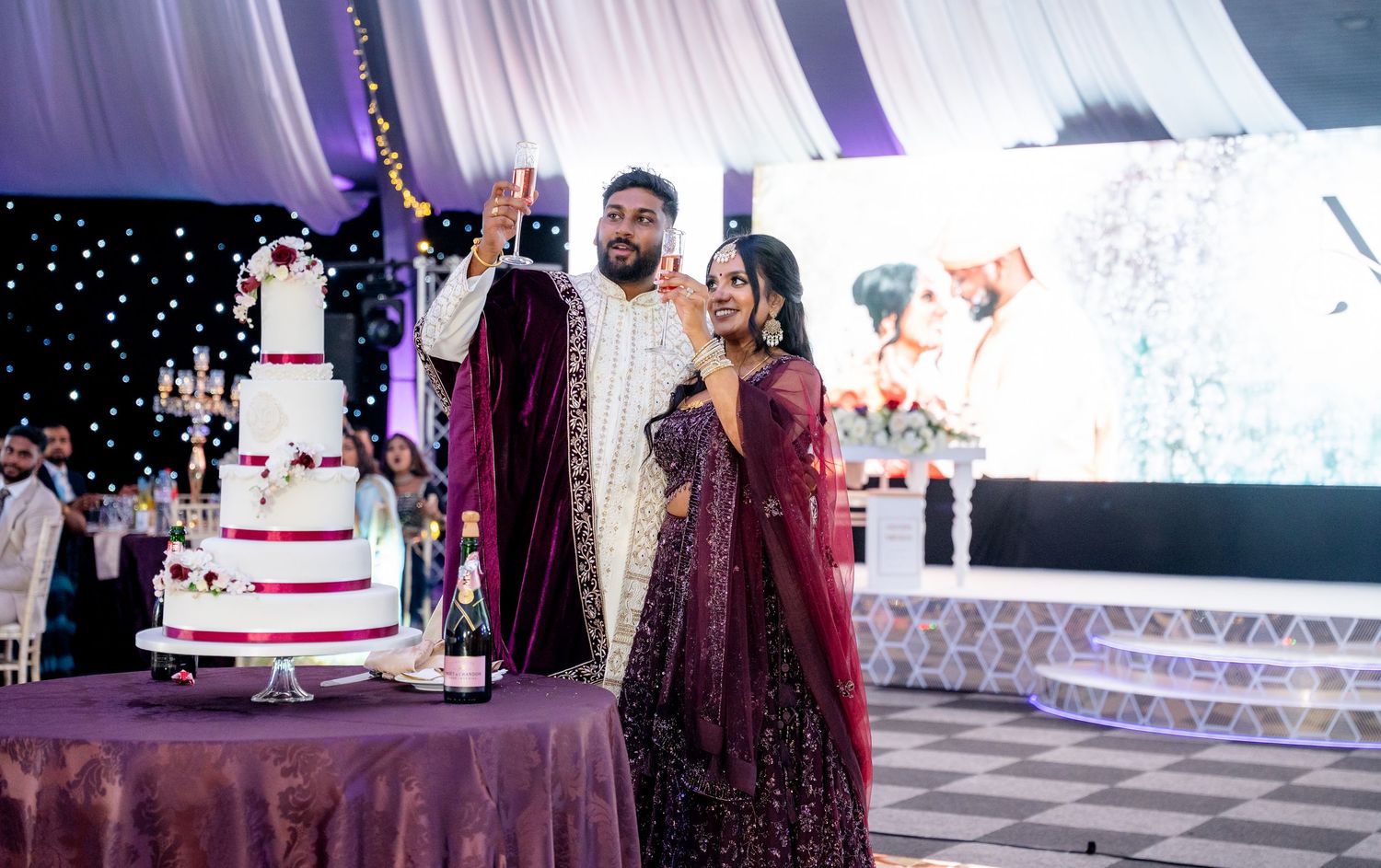 Bride and groom in traditional South Asian wedding attire cutting a white tiered cake at elegant reception.